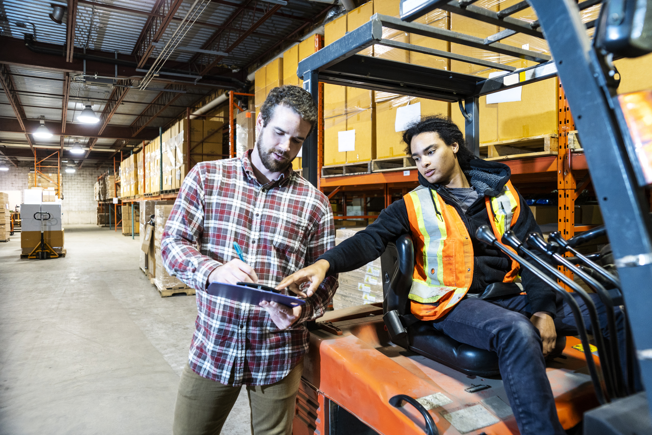 Two coworkers discussing in a warehouse.  One man has a clipboard and the other is on a forklift.