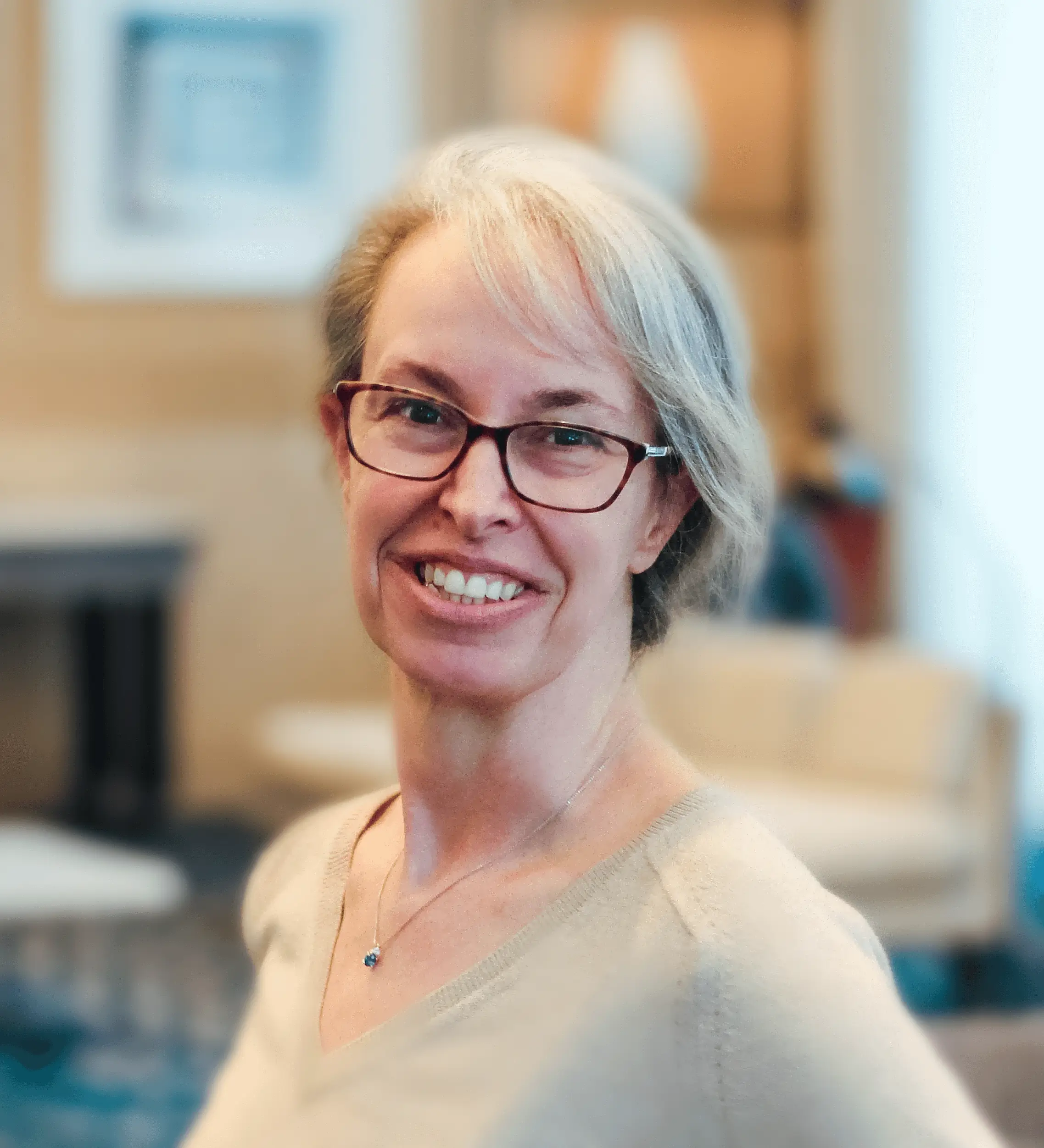 A woman with glasses and gray hair smiles at the camera in a warmly lit indoor setting with blurred furniture in the background.