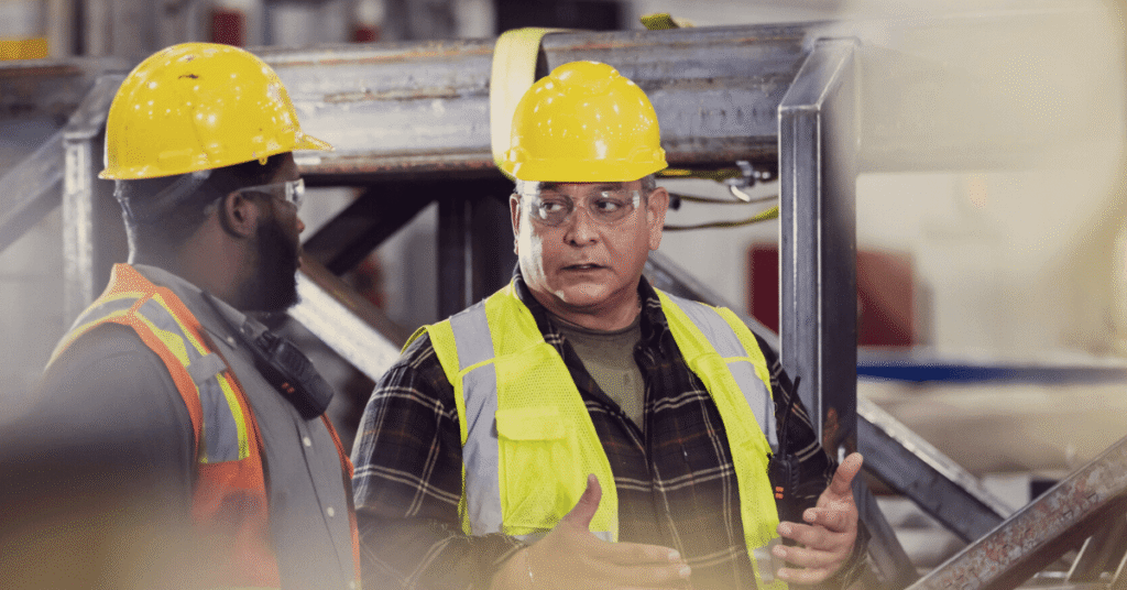 Two construction workers wearing yellow hard hats and safety vests discuss an automated workflows report from Silvi at a worksite with metal beams in the background.