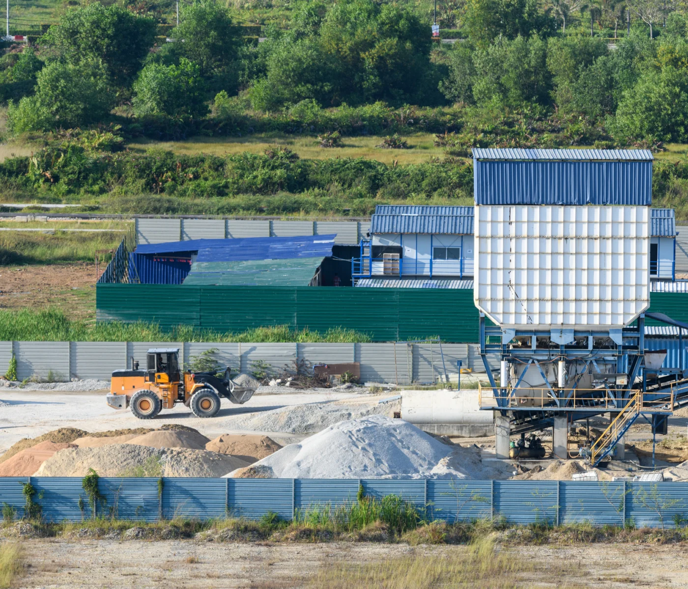 An industrial site with a yellow bulldozer moving sand and gravel piles, large machinery, and fenced buildings in the background.