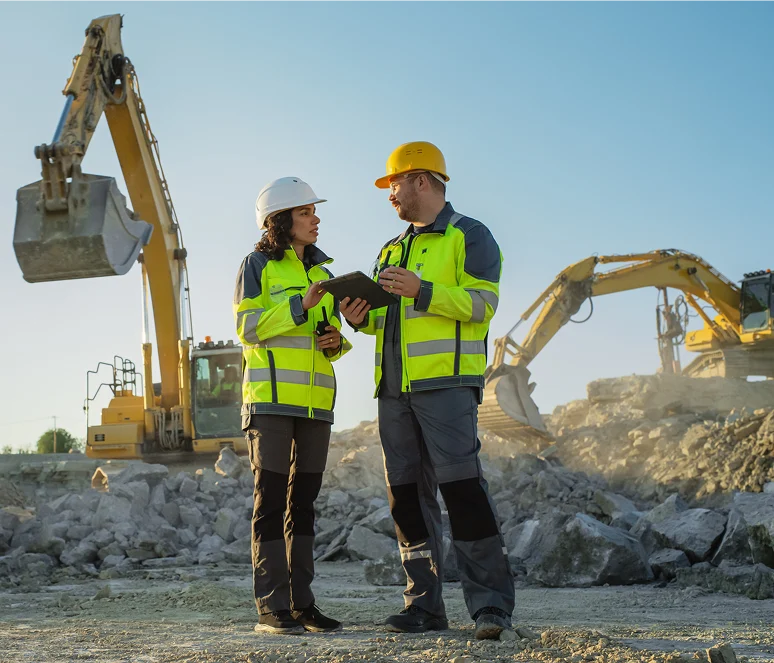 Two construction workers in safety gear stand on a rocky site, discussing a tablet, with excavators operating in the background under a clear sky.