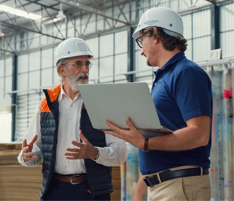 Two men wearing safety helmets discuss something in a warehouse; one holds a laptop while the other gestures with his hands.