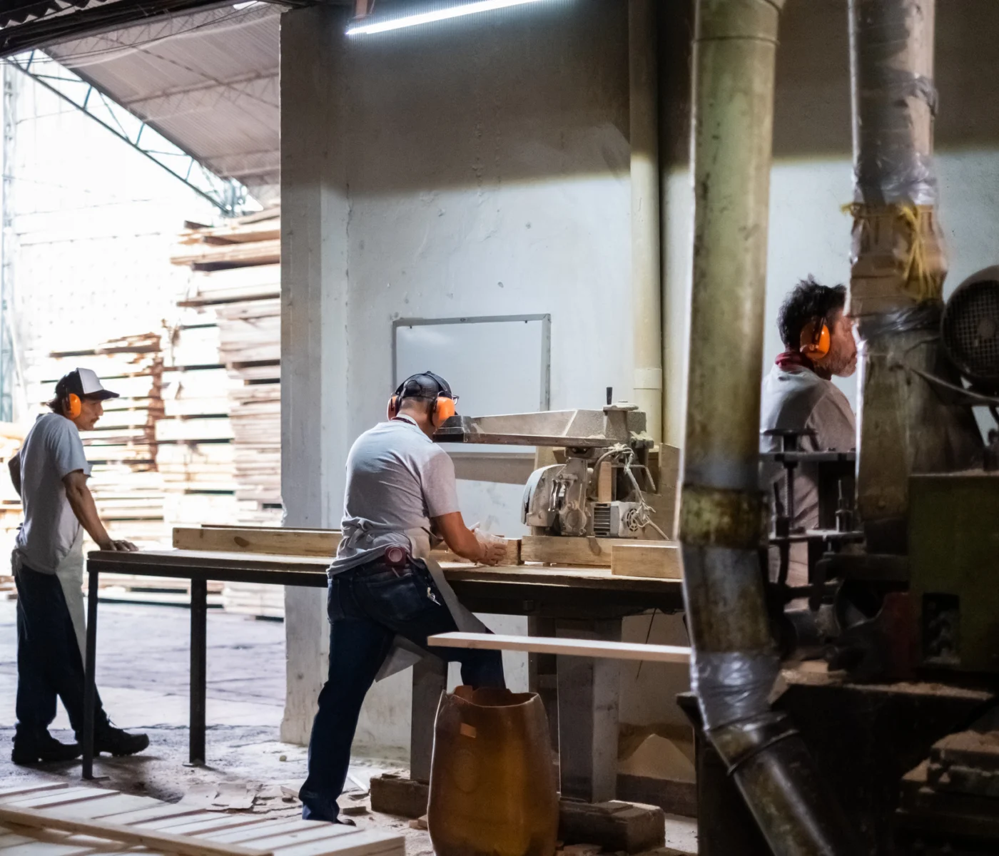 Three workers operate machinery and handle wood in a lumber mill or woodworking factory, surrounded by stacks of timber and industrial equipment.