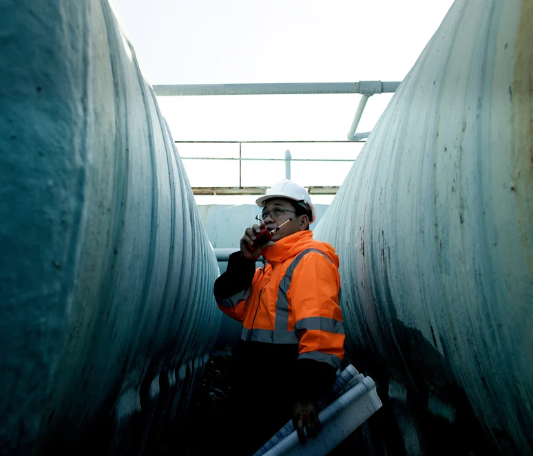 A worker in a hard hat and orange safety jacket stands between large industrial pipes, holding rolled-up plans and speaking into a two-way radio.
