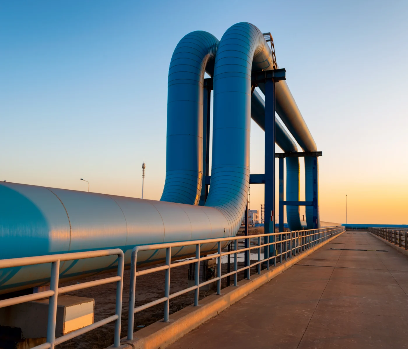 Large blue industrial pipeline structure on an elevated platform at sunset, with safety railings on both sides and a clear sky in the background.