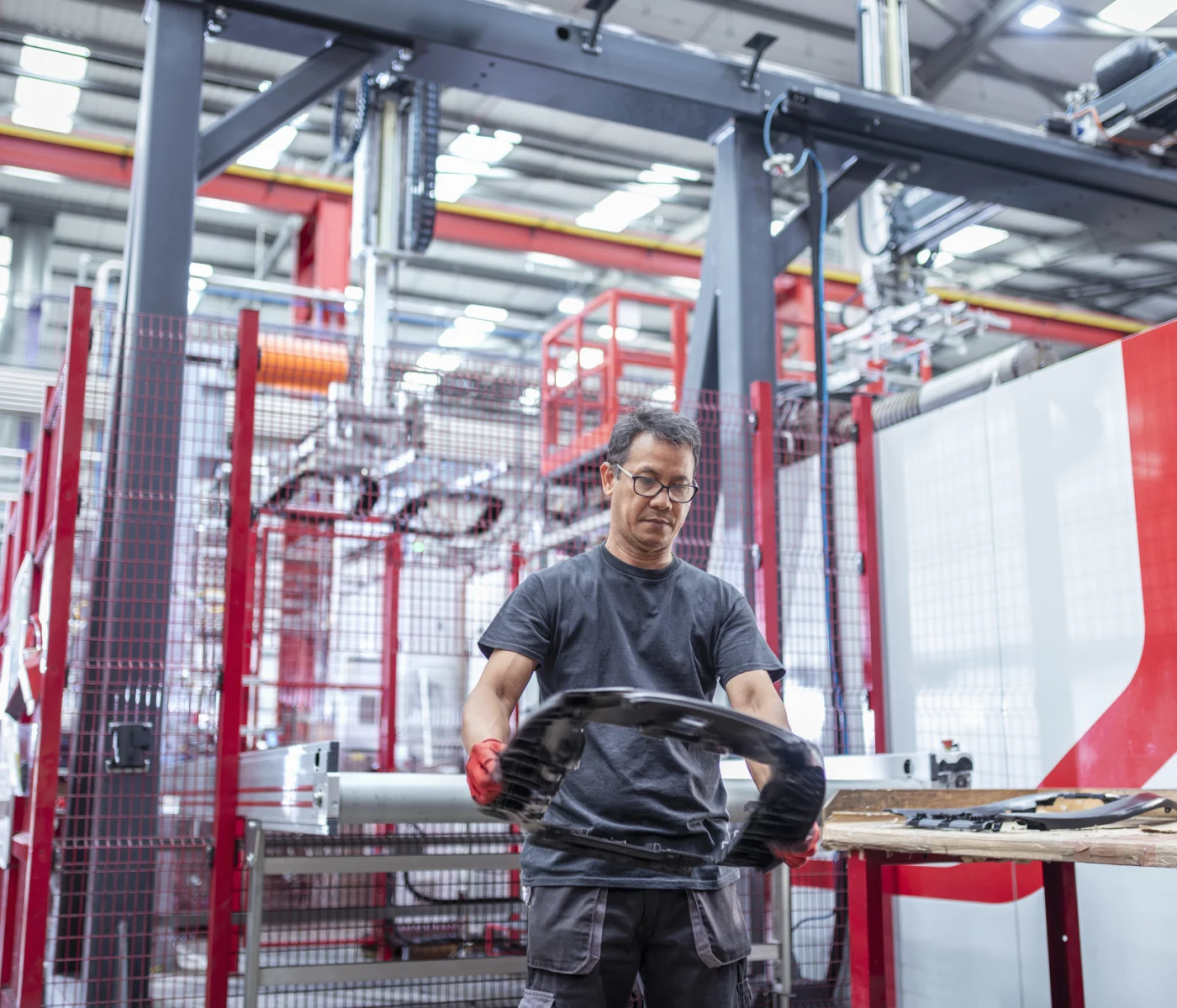 A man wearing gloves inspects a large metal part in a modern industrial factory with machinery and equipment in the background.