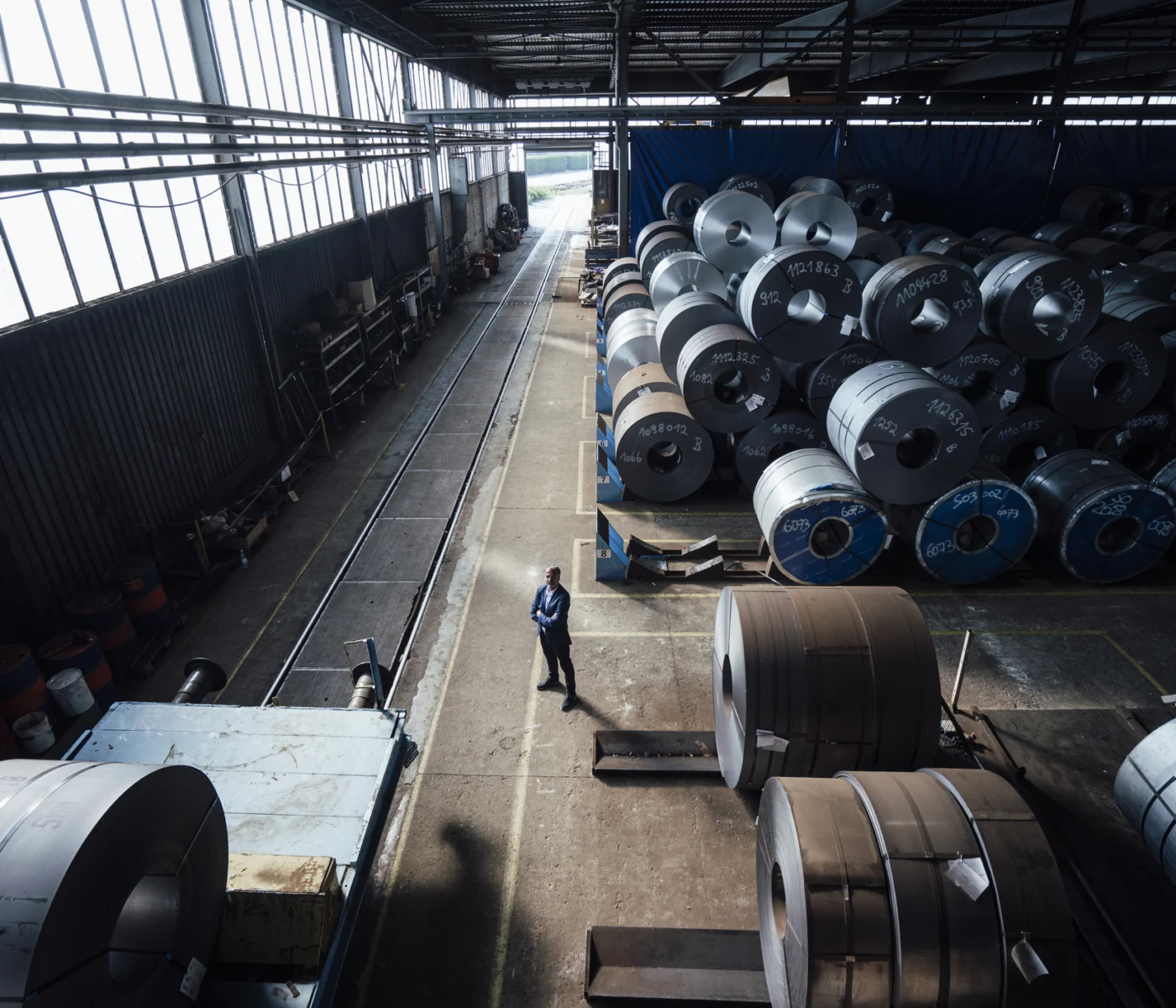 A person stands in a large industrial warehouse surrounded by numerous stacked metal coils and machinery, with natural light streaming through the windows.
