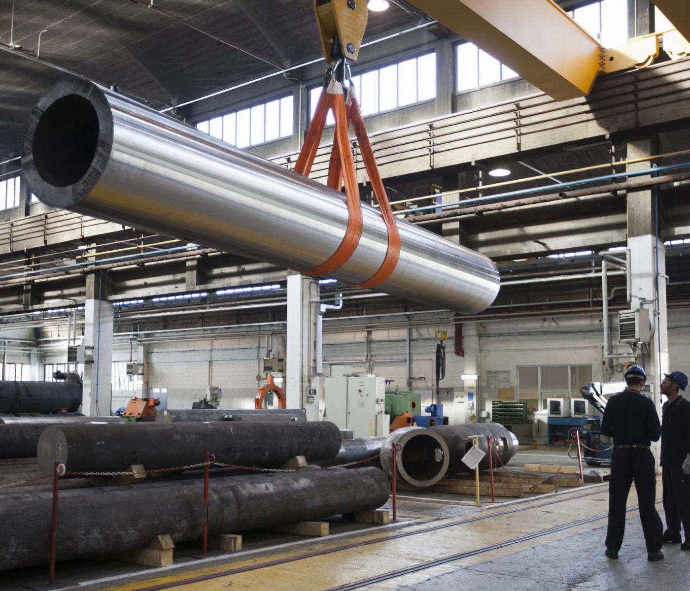 A large metal pipe is being lifted by an overhead crane in an industrial warehouse while two workers observe.