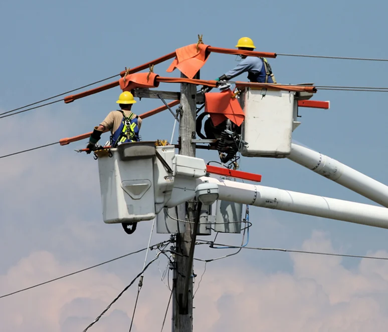 Two utility workers in bucket trucks repair or maintain power lines on a utility pole, wearing safety gear and helmets.
