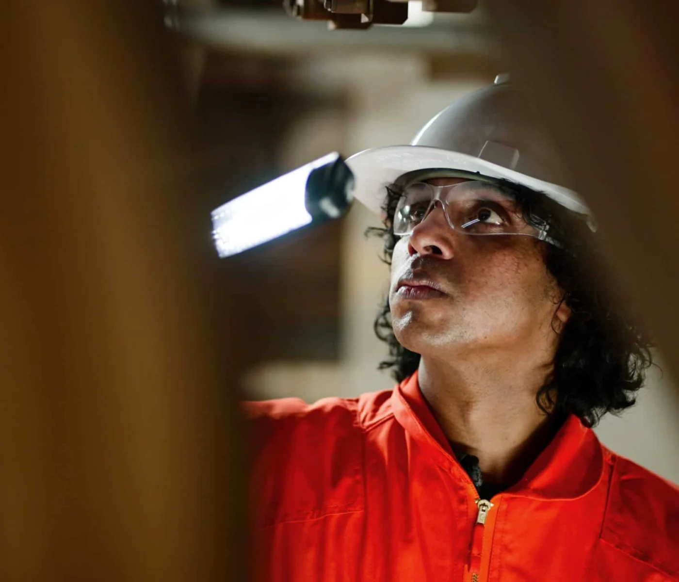 A worker in an orange safety suit and white hard hat inspects equipment indoors, with a focused expression and safety goggles on.
