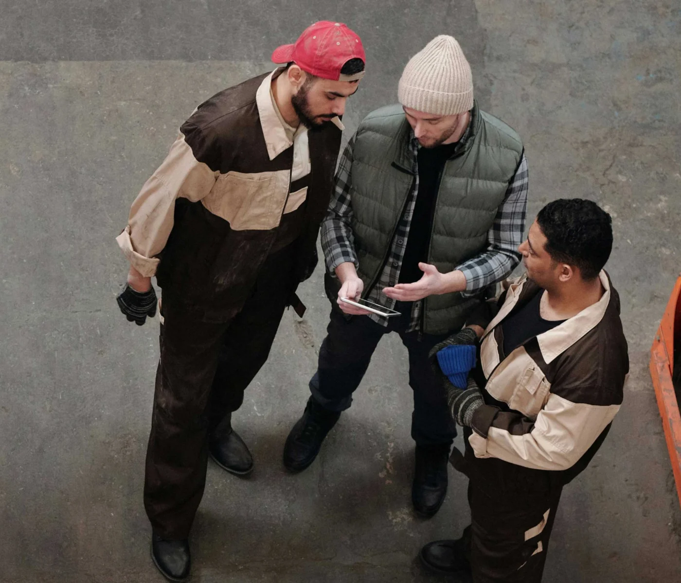 Three men in workwear stand together indoors, looking at a mobile device held by one of them.