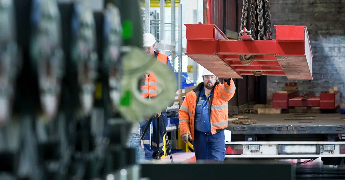 A worker in a high-visibility jacket, following strict safety programs, guides a large red metal object being lifted by a crane inside an industrial facility.
