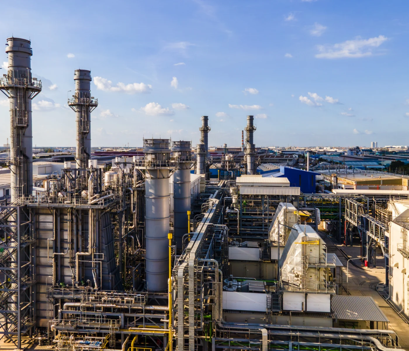 A large industrial plant with multiple smokestacks, metal structures, and pipelines under a blue sky with scattered clouds.