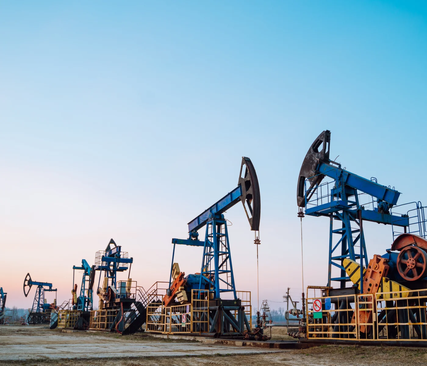 Several oil pumpjacks operate in a row on an oil field under a clear sky at sunset.