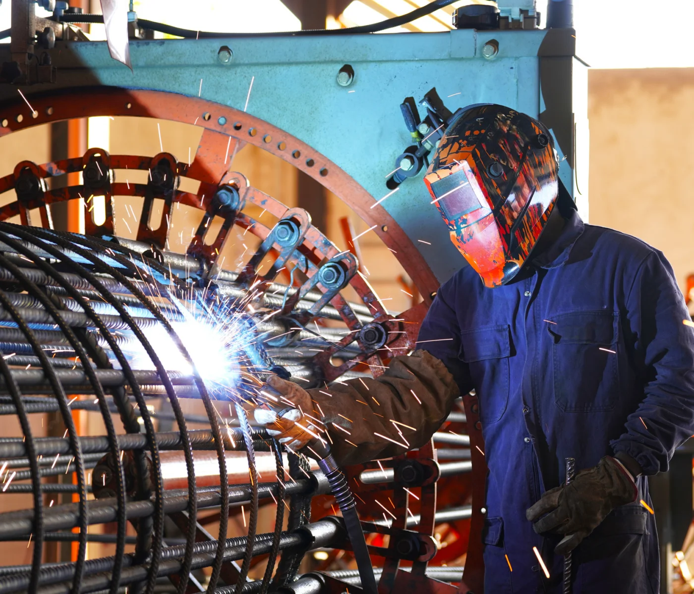 A worker in protective gear welds metal rods inside a large cylindrical structure at an industrial facility, with sparks flying.