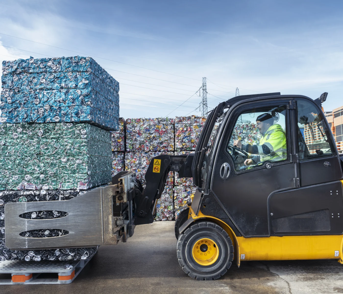 A forklift operator moves large compacted bales of crushed metal cans at a recycling facility, with stacks of similar bales in the background.