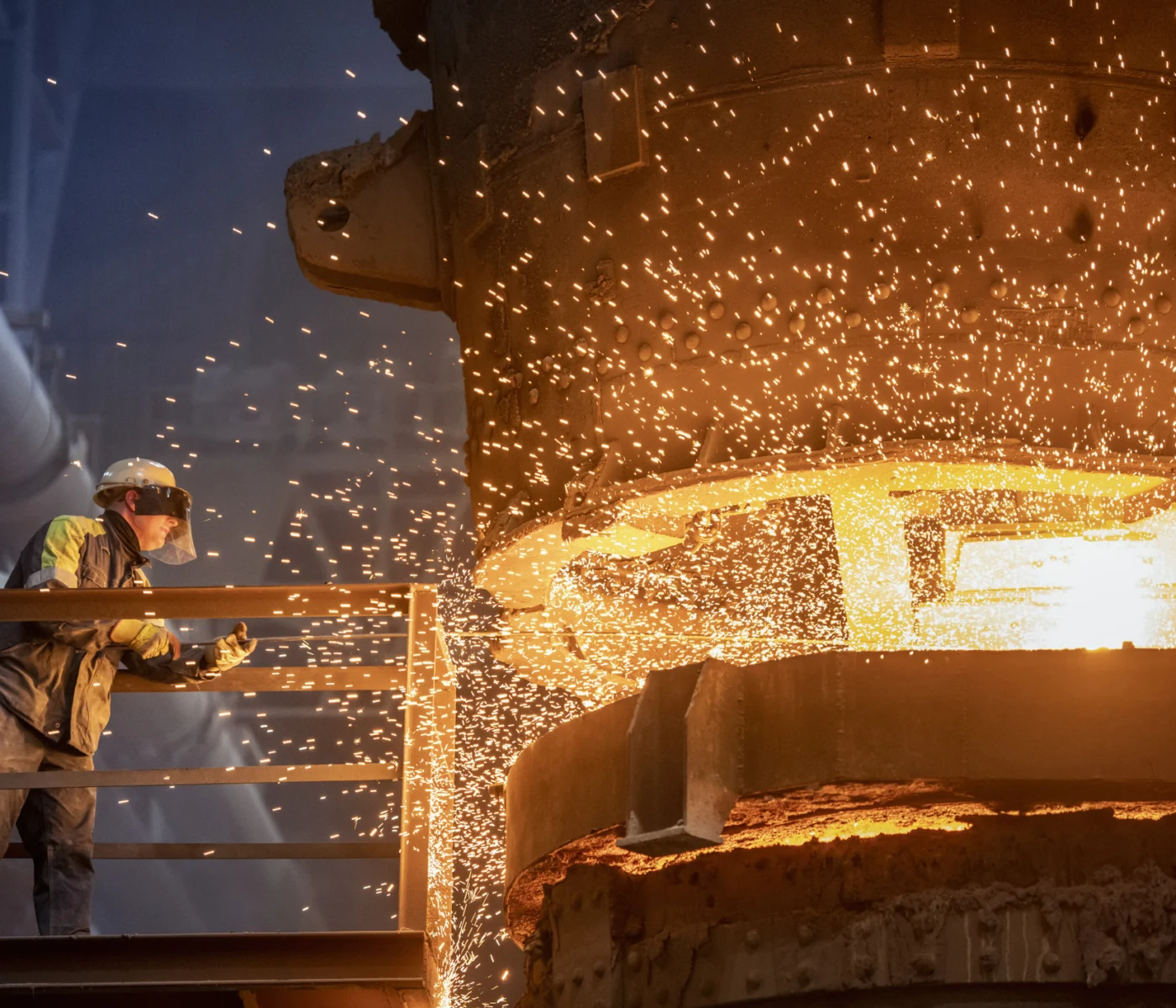 A worker in protective gear monitors molten metal pouring from a furnace, with bright sparks flying in an industrial setting.