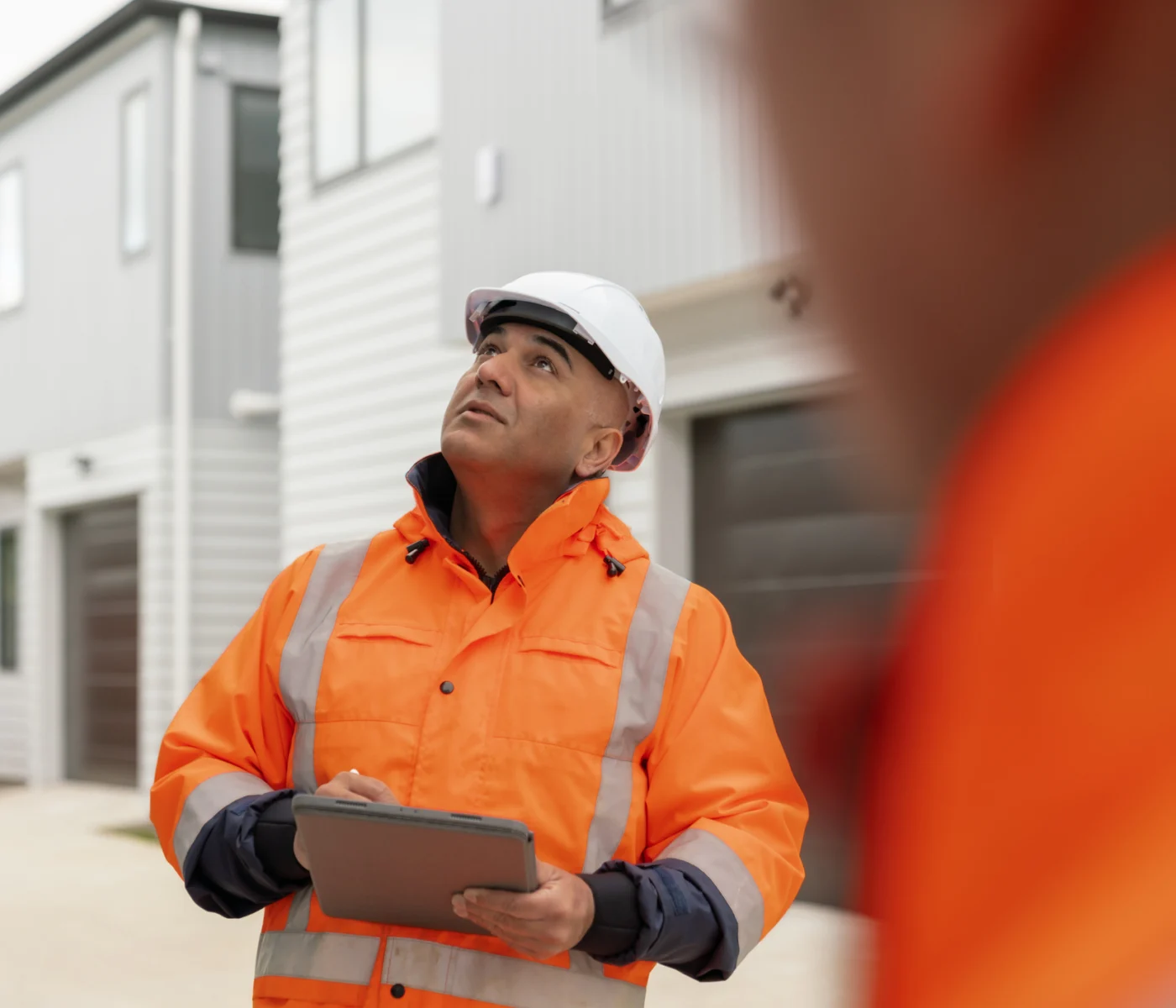 A construction worker in a safety helmet and orange jacket holds a clipboard while inspecting or observing a modern residential building outdoors.