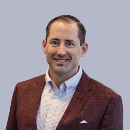 A man in a maroon blazer and checkered shirt smiles at the camera against a plain light gray background.