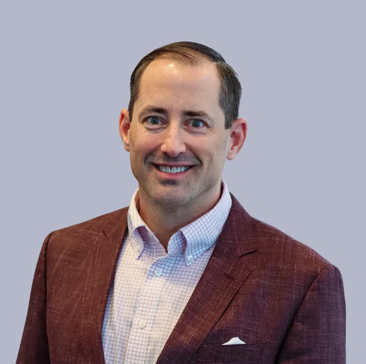 Man wearing a maroon blazer and light checkered shirt, smiling, posed against a plain light gray background.