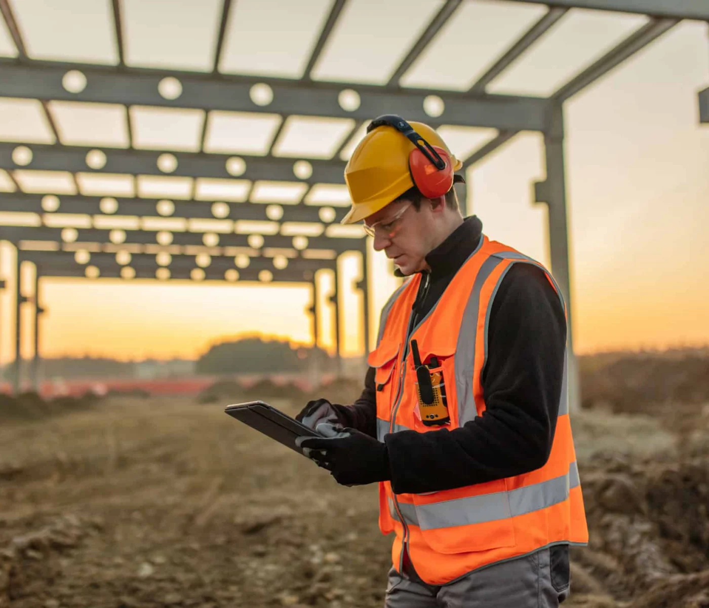 A construction worker in safety gear uses a tablet at a building site with a steel frame structure in the background at sunset.