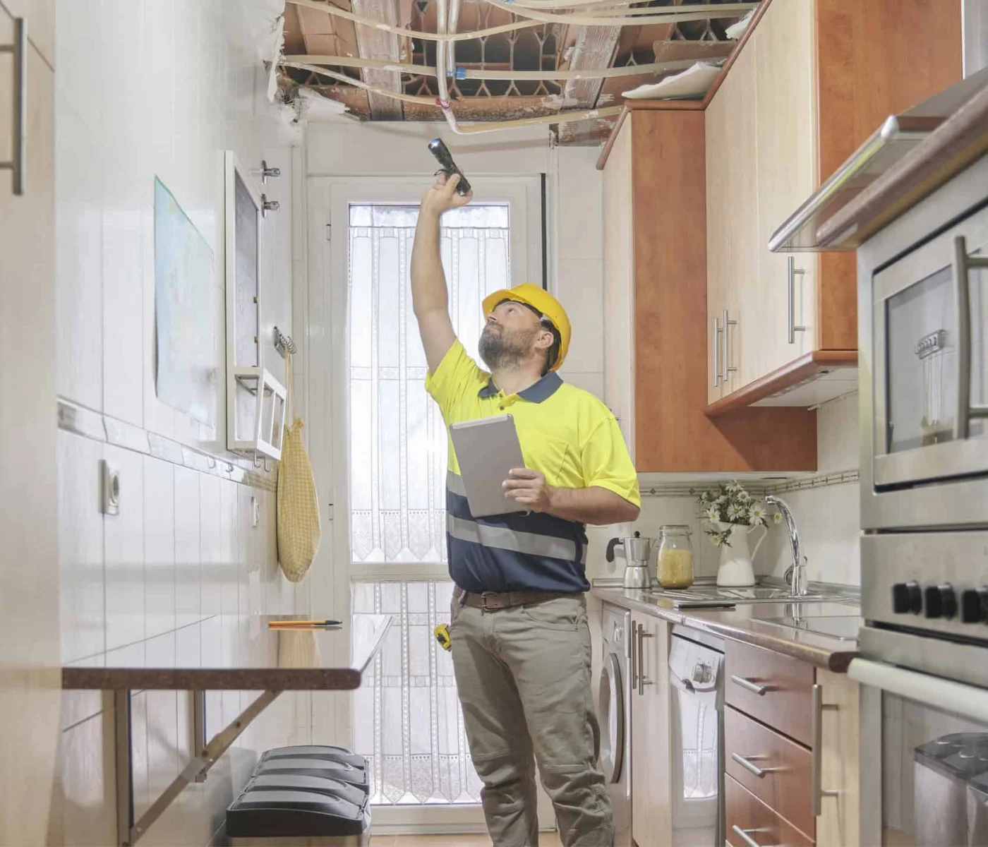 A construction worker wearing a hard hat inspects a kitchen ceiling with visible water damage and exposed pipes, holding a flashlight and a tablet.