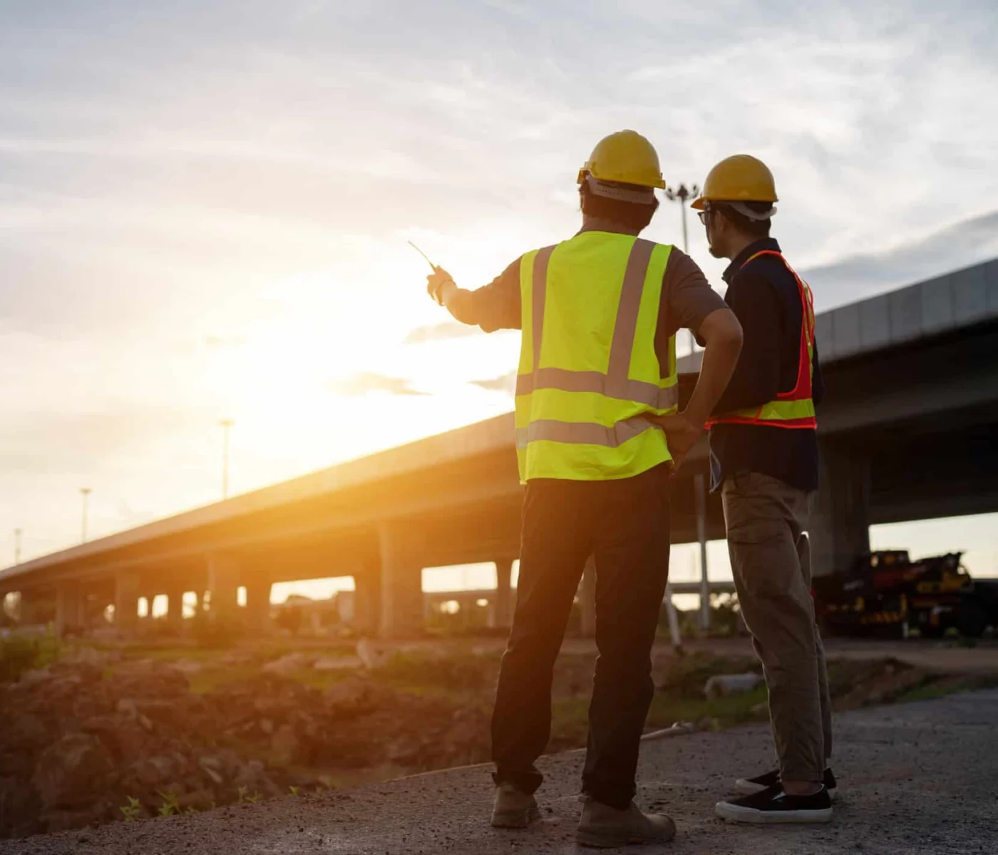 Two construction workers in safety vests and helmets stand near an overpass, one pointing towards the distance as they discuss the site at sunset.