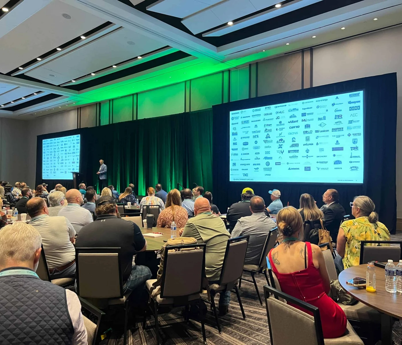A large group of people sit at round tables in a conference room, listening to a speaker on stage. A large screen displays numerous company logos.