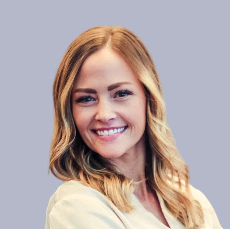 A woman with long, wavy blonde hair and light skin smiles at the camera against a plain light gray background.