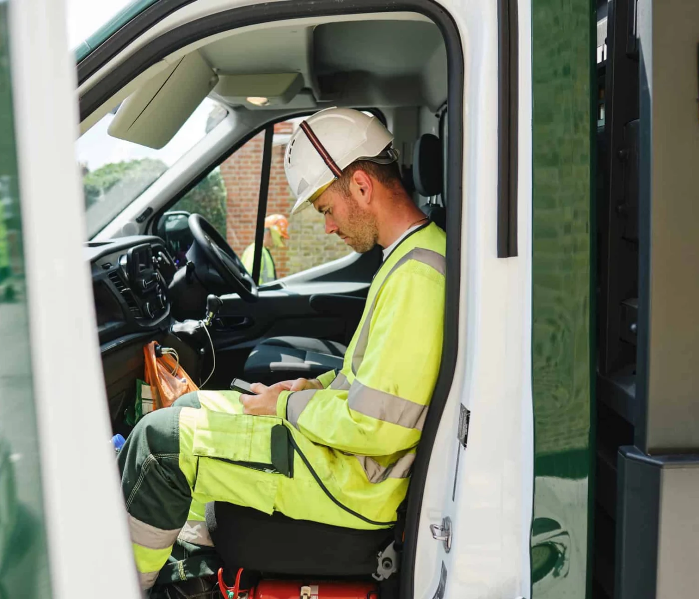 A construction worker in a high-visibility suit and hard hat sits inside a parked work van, looking down at something in his hands.