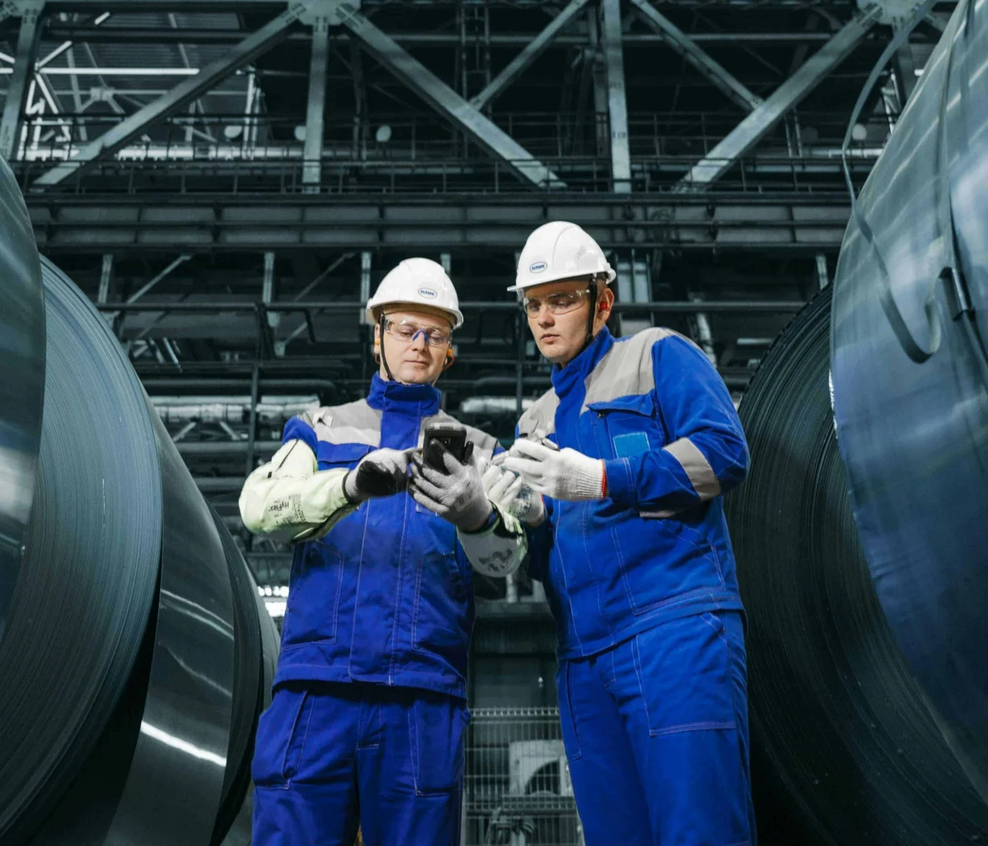 Two workers in blue uniforms and helmets examine a device while standing between large metal coils in an industrial facility.