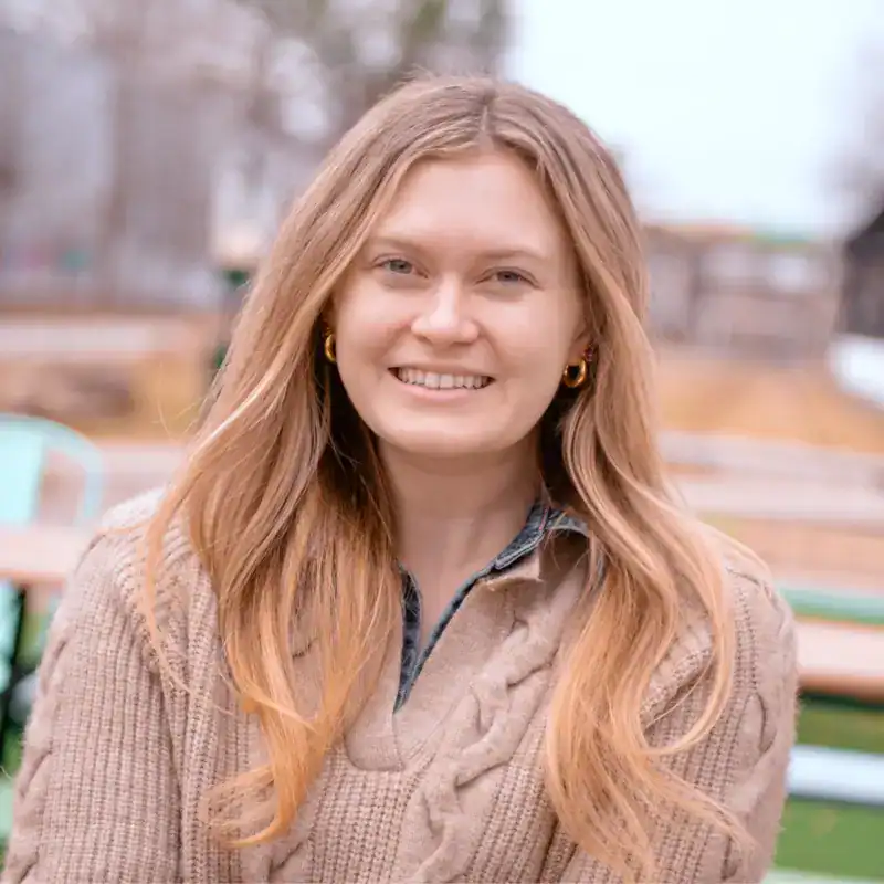 Caroline Miller, with long blonde hair, wears a tan cable-knit sweater and gold hoop earrings as she sits outdoors, smiling at the camera—reflecting her warm approach to data-driven decision-making.