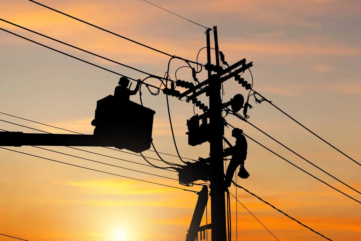Two utility workers repair electrical lines on a pole at sunset; one is in a bucket lift, and the other is harnessed to the pole.