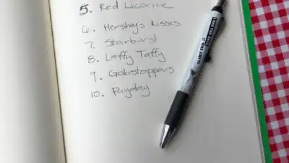 Open notebook on a red checkered table with a list of ten candies written in black ink and a pen resting on the page.