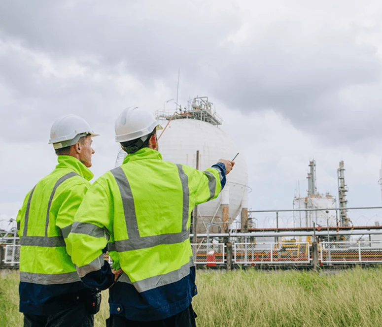 Two workers in high-visibility jackets and hard hats stand outdoors, one pointing toward industrial gas storage tanks at a facility.