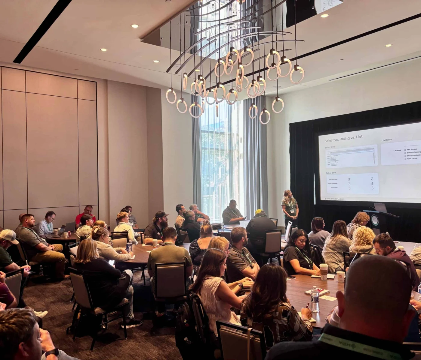 A group of people sit in a conference room watching a presentation projected on a screen at the front of the room.