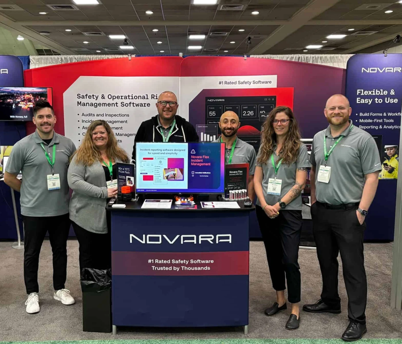 Six people stand and smile at a trade show booth for Novara, featuring banners about safety and operational risk management software and a display screen with promotional material.