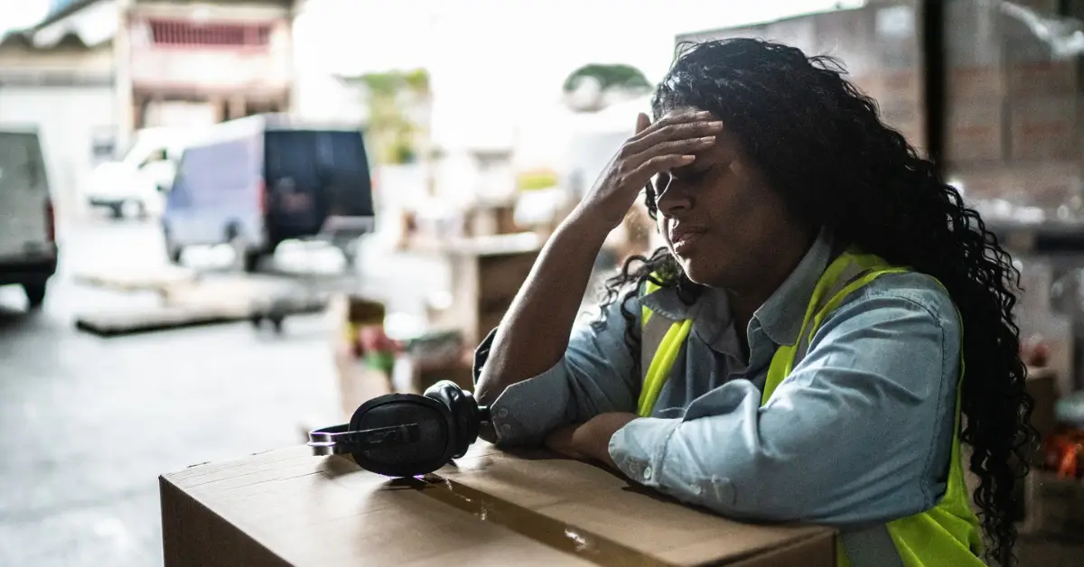 A worker in a safety vest leans on a box with a tired expression, resting her head in her hand, highlighting the importance of heat illness prevention and field safety in the busy Gallo Mechanical warehouse setting.