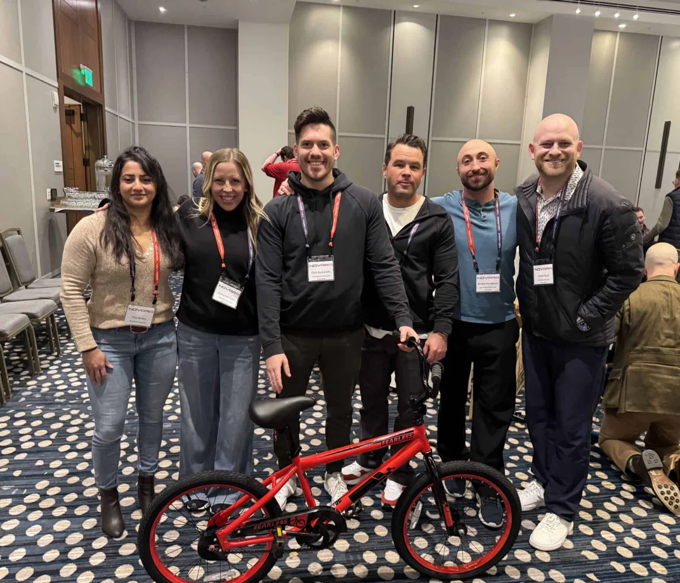 Six adults stand indoors behind a red children's bicycle, all wearing conference name badges. The setting appears to be a conference room with chairs and patterned carpet.