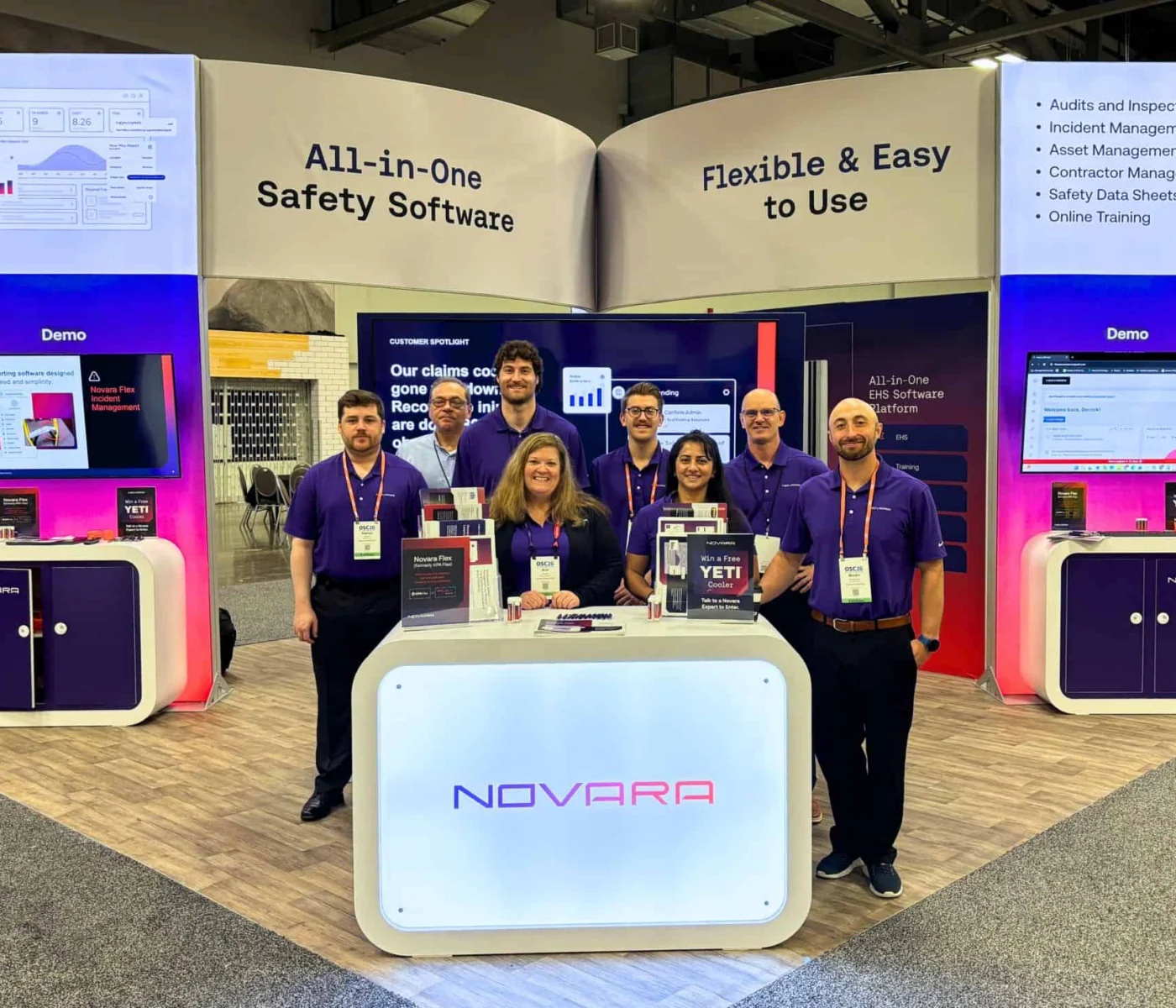 Seven people stand at a trade show booth for Novara, an all-in-one safety software, with screens displaying demos and informational banners in the background.