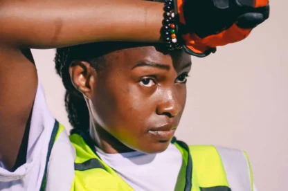 A person wearing a neon yellow safety vest and orange glove wipes sweat from their forehead, looking tired.