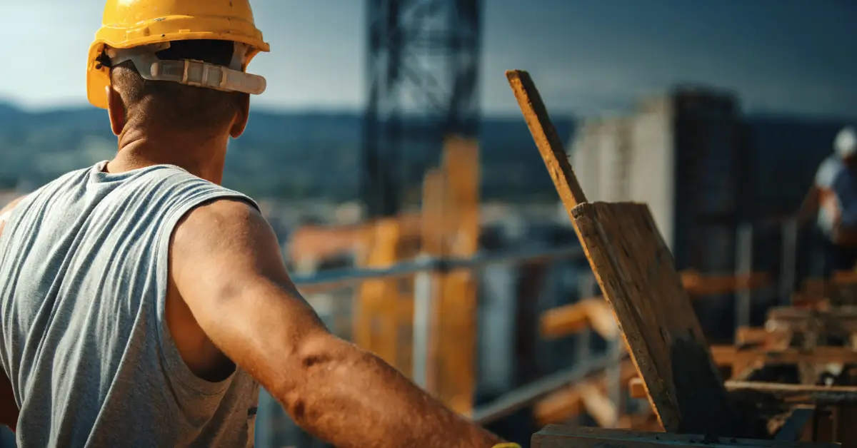 Construction worker in a yellow hard hat and gray shirt stands at a building site, looking toward unfinished structures under a clear sky during an unannounced inspection.