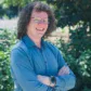 Person with curly hair, glasses, and a blue shirt stands outdoors with arms crossed, smiling in front of green foliage and red berries.
