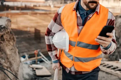 Man in an orange safety vest holding a white hard hat and a phone at a construction site.