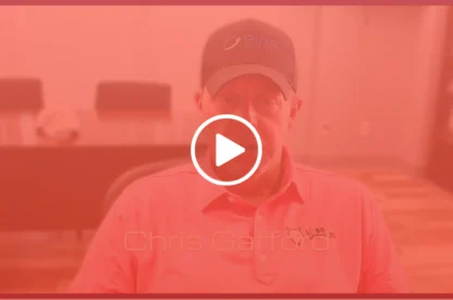 A man wearing a baseball cap and a polo shirt sits in an office; video play button overlay.