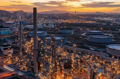 Aerial view of an industrial oil refinery complex at sunset, with numerous storage tanks and glowing lights throughout the facility.