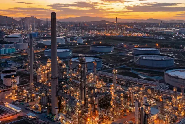 Aerial view of an industrial oil refinery complex at sunset, with numerous storage tanks and glowing lights throughout the facility.
