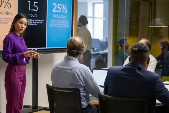 A woman in a purple outfit presents data on task-switching efficiency to a group of colleagues in a conference room.